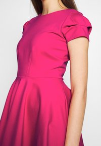 Femme portant une robe ajustée rose vif avec des manches courtes bouffantes et une jupe évasée, montrée des épaules à la taille.