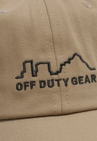 Beżowa czapka materiałowa z czarnym wyszywanym logo przedstawiającym panoramę miasta przechodzącą w szczyty gór oraz z napisem "OFF DUTY GEAR."