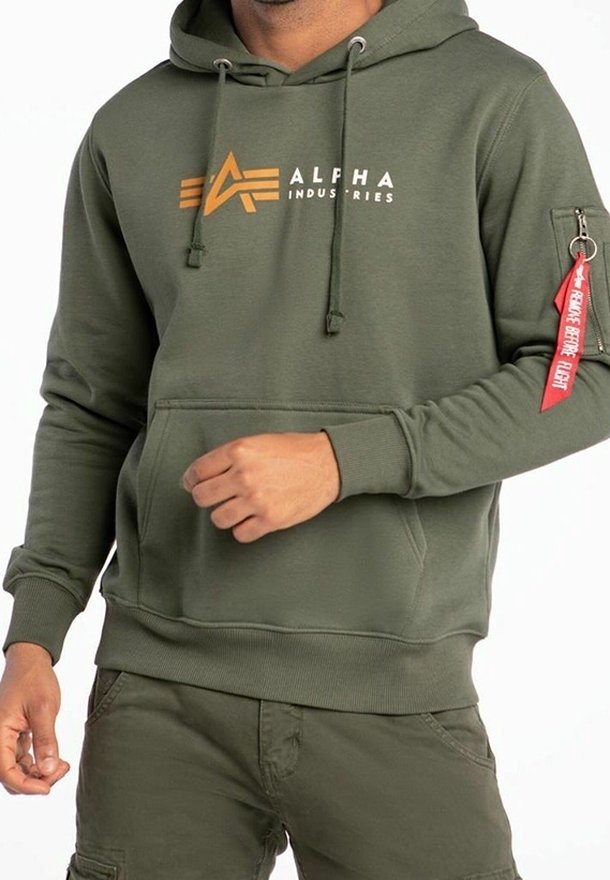 Olive green hoodie with a kangaroo pocket, drawstrings, large "ALPHA INDUSTRIES" logo in orange, and a side pocket with a red tag.