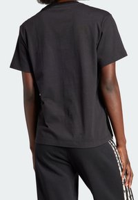 Person som bär en enkel svart kortärmad tröja och svarta byxor med beige leopardmönstrade sidostreck, sedd bakifrån.