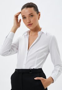 Biała koszula z guzikami, z kołnierzykiem i długimi rękawami, o dopasowanym kroju i gładkiej fakturze, zestawiona z czarnymi spodniami.