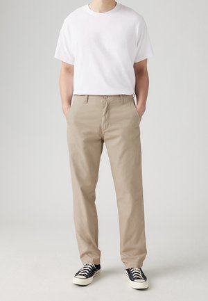 Mann, der ein schlichtes weißes kurzärmeliges T-Shirt, beige gerade geschnittene Hose und schwarze niedrige Sneaker mit weißen Schnürsenkeln trägt.