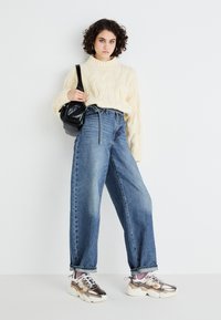 Cream cable-knit sweater, high-waisted blue denim wide-leg jeans, and metallic-accented sneakers. Black crossbody bag. White background.