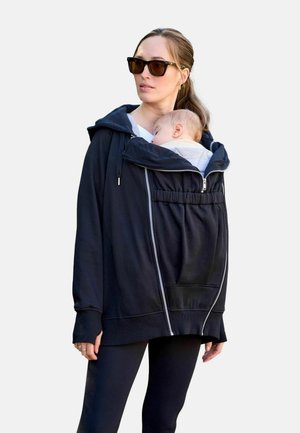 Kvinde iført solbriller bærer en baby, der sover inde i en sort lynlåstrøje til bæresele med hætte og lommer.