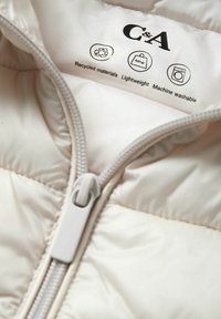 Doudoune beige légère avec fermeture éclair à l'avant, fabriquée à partir de matériaux recyclés. Les étiquettes d'entretien indiquent qu'elle est lavable en machine et légère.