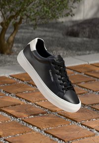 Svart lær sneakers med en hvit hælaksent, rund tå og svarte snøring. Har en logo på siden og en teksturert gummisåle.