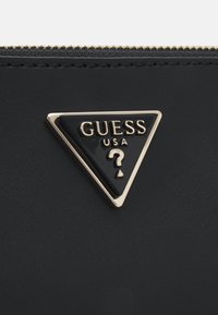 Černá peněženka s trojúhelníkovým zlatým kovovým detailem s embosovaným nápisem "GUESS USA" a otazníkem, umístěná na texturovaném pozadí.