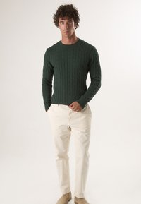 Zielony sweter z dzianiny w sploty, z okrągłym dekoltem, w zestawie z kremowymi spodniami. Sweter ma teksturowe wzory i ściągacze przy rękawach.