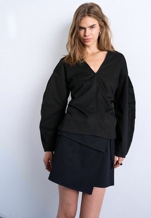 Ung kvinde med løst lys brunt hår iført sort V-hals langærmet top og sort asymmetrisk nederdel, stående mod en hvid væg.