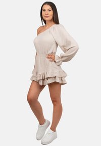 Beige einseitig trägervarianter Kleid mit strukturiertem Stoff, Rüschen-Saum und langen Ärmeln. Kombiniert mit weißen Turnschuhen für einen lässigen Look.