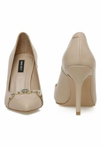 Beiga högklackade skor med spetsig tå, glänsande yta och en dekorativ guldsmyckeskedja fram. Stilettklackdesign.