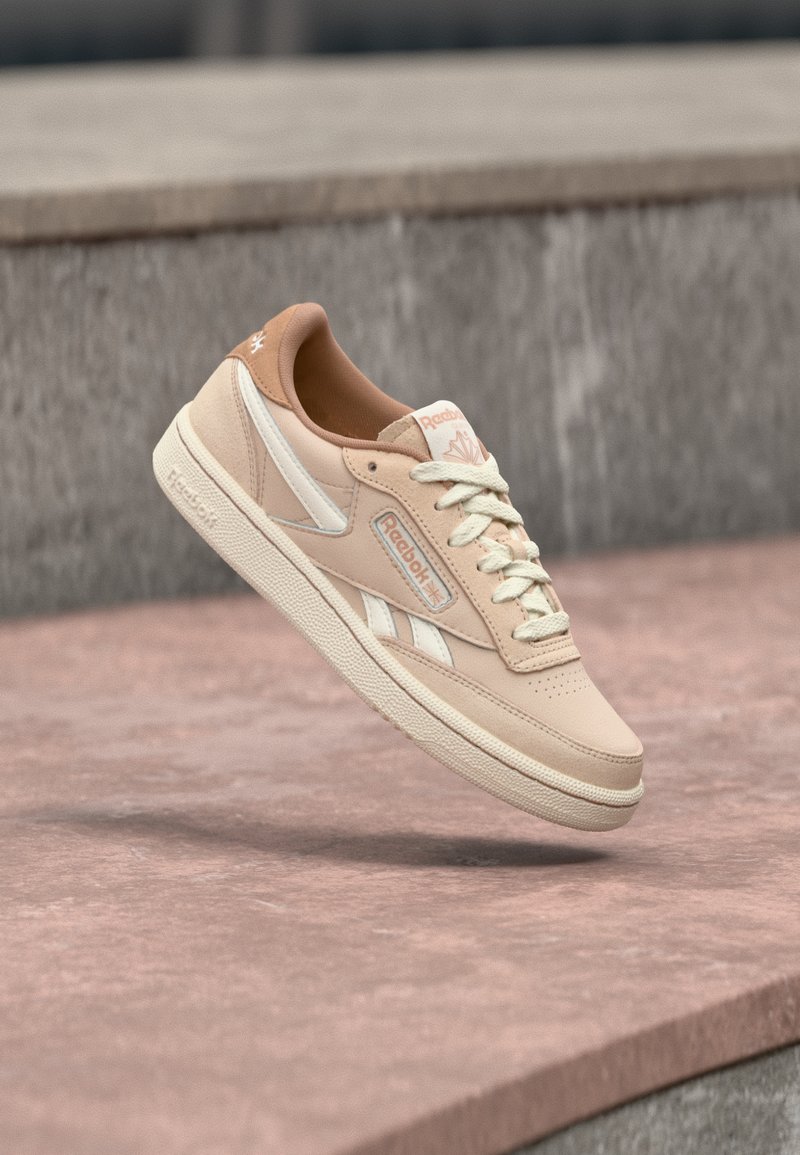 Sneaker Reebok beige e bianco con lacci galleggianti sopra una superficie di pietra rossastra e uno sfondo in cemento grigio.