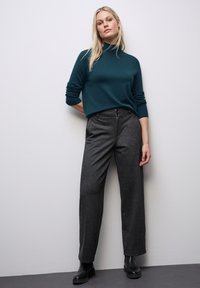 Teal coltrui gecombineerd met hoge taille, wijde charcoal broek. Model staat met handen op de heupen, draagt zwarte enkellaarsjes.