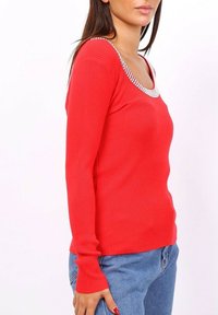 Pull long à manches longues rouge côtelé avec un large col en forme de scoop orné d'un accent en argent. Le tissu est texturé et ajusté.