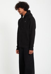 Black zip-up hoodie with a front pocket, drawstring hood, soft texture, and a small logo accent on the chest. Worn with loose black pants.