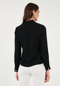 Black long-sleeve blouse made of smooth fabric, featuring a high neckline, back zipper, and contrasting button accents at the cuffs.