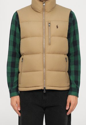 Beige wattierte Weste mit Reißverschluss und Brusttasche, getragen über einem grün-schwarz karierten Langarmhemd und schwarzen Hosen.