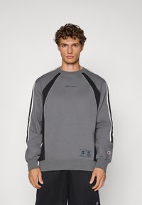 Sudadera gris con acentos negros, rayas blancas a lo largo de las mangas, puños acanalados y cuello redondo. Presenta un pequeño logo en el pecho.
