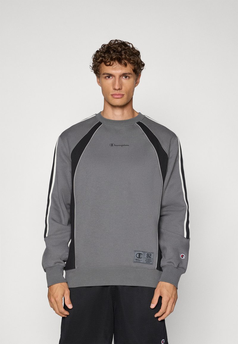 Sudadera gris con acentos negros, rayas blancas a lo largo de las mangas, puños acanalados y cuello redondo. Presenta un pequeño logo en el pecho.