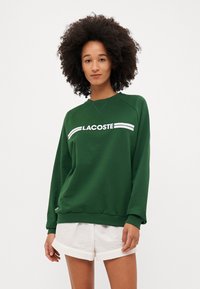 Hanorac verde cu logo-ul alb "LACOSTE" și dungi pe piept. Material moale, mansete cu jerseu și croială lejeră. Asortat cu pantaloni scurți albi.