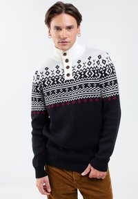 Kootud pullover-sviiter kõrge krae ja kreemisarnase ülaosaga, tumedast mereteest kehaga, millel on geomeetrilised mustrid mustas ja punases, aktsentidega pruuni nuppudega.