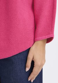 Fukszia kötött pulóver bordázott mintával és háromnegyedes ujjakkal, sötét farmernadrággal párosítva; kéz pihentetve a farmerral.