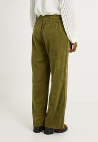 Pantalones de pana verde oliva con un diseño de pierna ancha, cinturilla elástica y textura de ribetes verticales. Calzado marrón visible en la parte inferior.