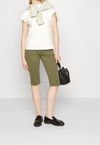 Žena v bílé krátké košili s lehkým svetrem přes ramena, olivově zelených kalhotách ke kolenům, černých mokasínech, držící malou černou kabelku.