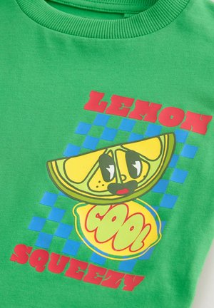 T-shirt di cotone verde con grafica di un limone, testo rosso "LEMON" e "SQUEEZY", e motivo di sfondo a quadretti blu.