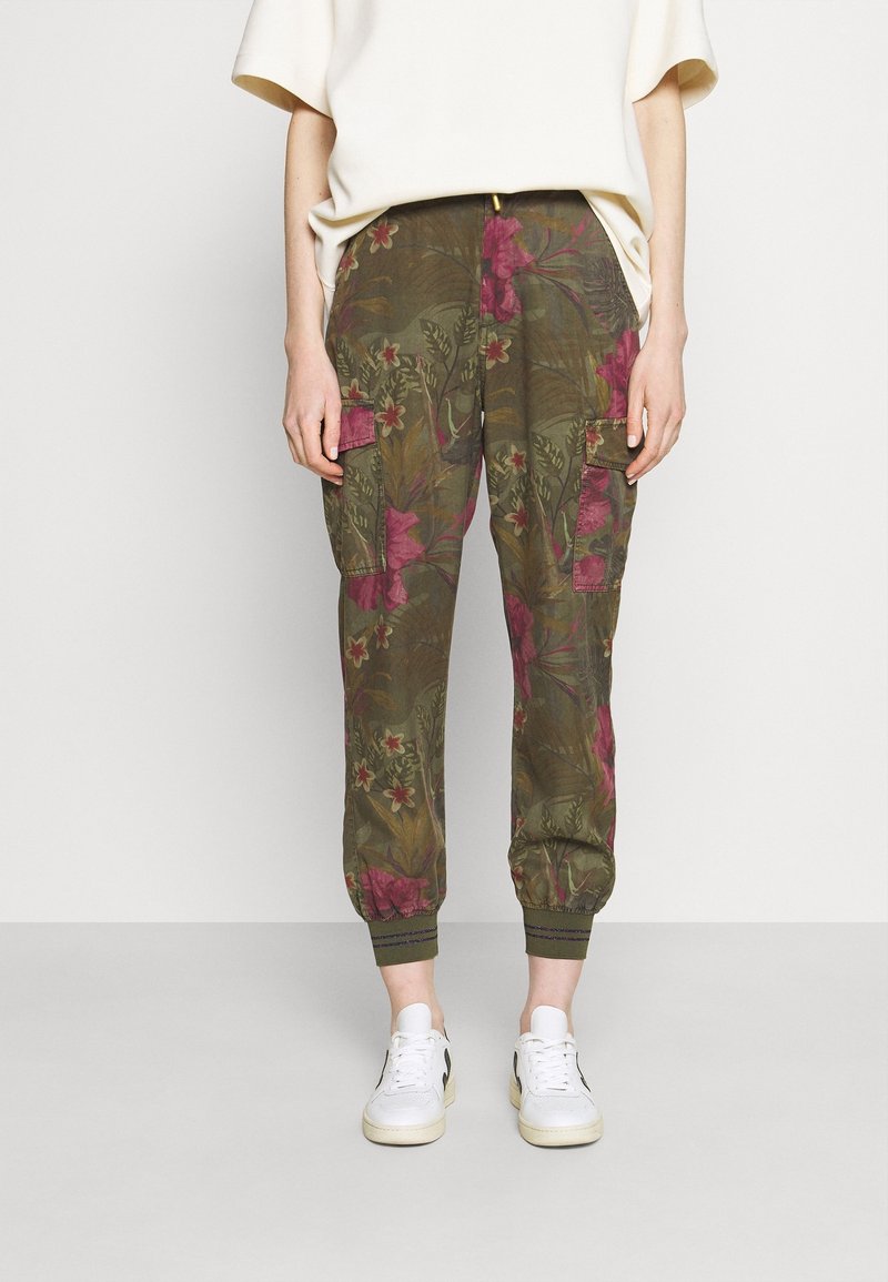 Personne portant un pantalon cargo vert olive avec imprimé floral rose, poignets élastiques, associé à des baskets blanches et un haut blanc ample.