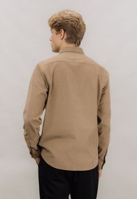 Brun langermet skjorte med klassisk krage, knappmanchetter og et teksturert stoff, vist fra baksiden mot en nøytral bakgrunn.
