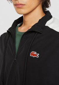 Svart lättviktig jacka med hög krage, framzipper och kontrasterande vita axelpaneler. Har en röd broderad krokodil-logotyp.