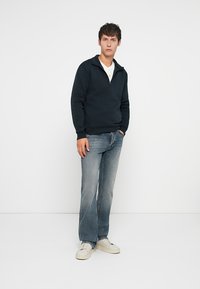 Sweat-shirt zippé bleu marine avec un col montant, associé à un jean bleu usé et des baskets blanches. Tissu lisse, coupe décontractée et design minimaliste.