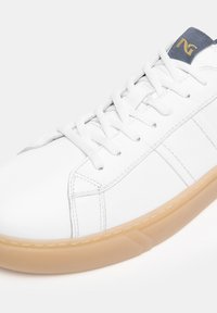 Weißer Sneaker mit strukturiertem Leder-Obermaterial, flachen weißen Schnürsenkeln und einer Sohle aus Gummi. Merkmale umfassen einen marineblauen Akzent auf der Zunge mit geprägtem Logo.