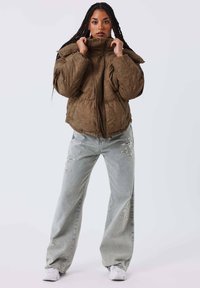 Chaqueta acolchada marrón con cuello alto y cierre de cremallera. Se lleva sobre unos vaqueros desgastados azul claro. Zapatillas blancas completan el conjunto.