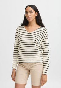 Gestreifter Pullover in Creme und Schwarz, mit V-Ausschnitt, langen Ärmeln und einem strukturierten Stoff. Getragen mit beigen Shorts.