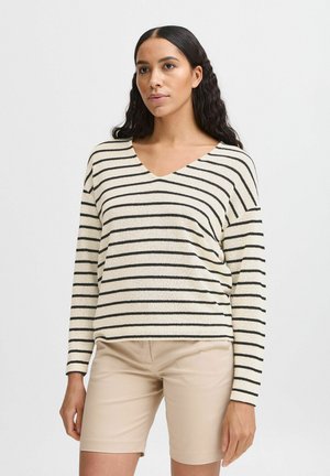 Gestreifter Pullover in Creme und Schwarz, mit V-Ausschnitt, langen Ärmeln und einem strukturierten Stoff. Getragen mit beigen Shorts.