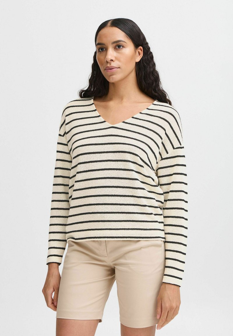 Gestreifter Pullover in Creme und Schwarz, mit V-Ausschnitt, langen Ärmeln und einem strukturierten Stoff. Getragen mit beigen Shorts.