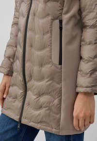 Chaqueta acolchada beige con un acabado mate y suave, que presenta un bolsillo con cremallera y detalles de cremallera negra a lo largo de las costuras frontales y laterales.