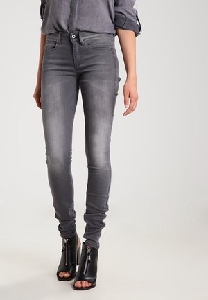 Femme portant un jean gris ajusté et délavé ainsi que des bottines noires ouvertes à l'avant avec des fermetures éclair frontales, debout devant un fond uni.