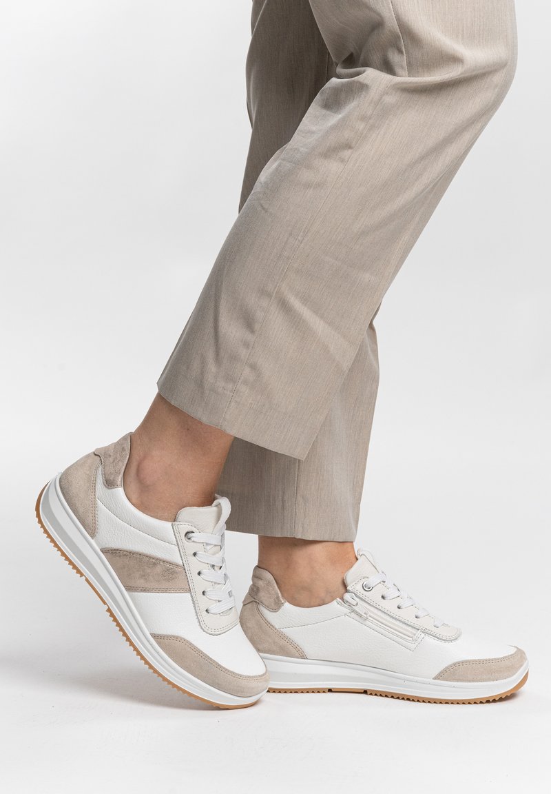 Person wearing white and beige lace-up sneakers with side zipper, standing on one foot, beige pants cropped above the ankle.