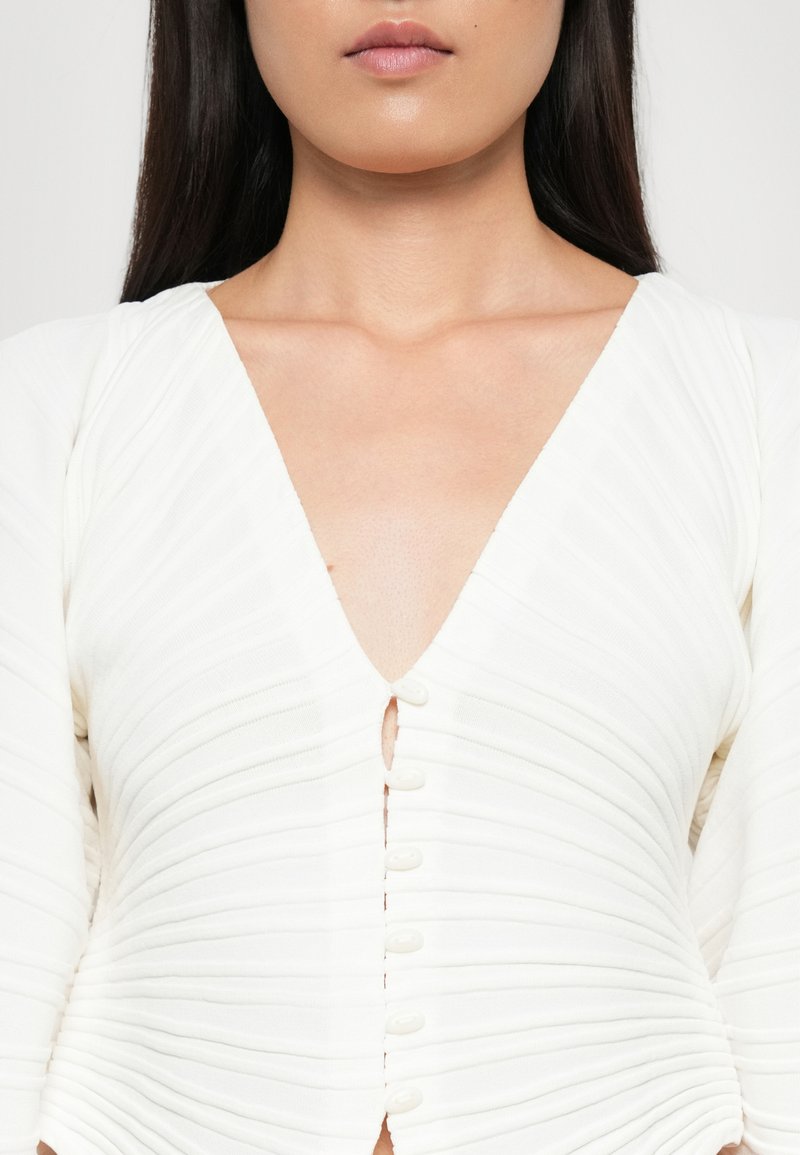 Haut blanc côtelé avec un décolleté en V profond, comportant des boutonnages sur le devant et de longues manches bouffantes. Texture lisse, design ajusté.