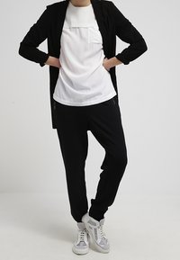 Giacca nera lunga, top bianco con tasca, joggers neri e sneakers bianche con dettagli grigi. L'outfit presenta un fit rilassato e un design a strati.