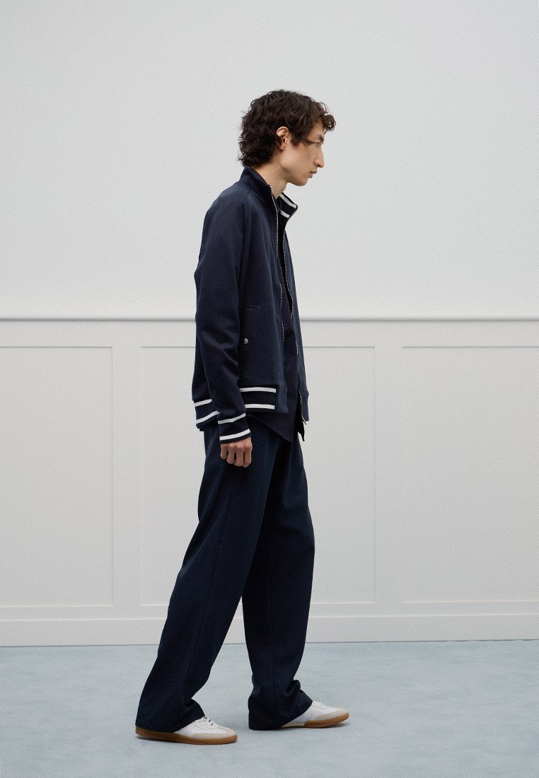 Granatowa kurtka z białymi prążkowanymi mankietami, nałożona na ciemną koszulę, zestawiona z szerokimi granatowymi spodniami i białymi trampkami. Gładka tekstura.