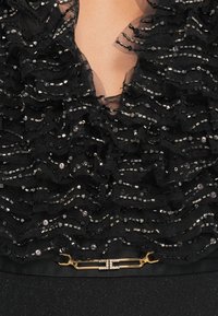 Top de tecido preto com camadas de babados adornadas com lantejoulas e contas, com um decote em V e um fecho de metal dourado na bainha.