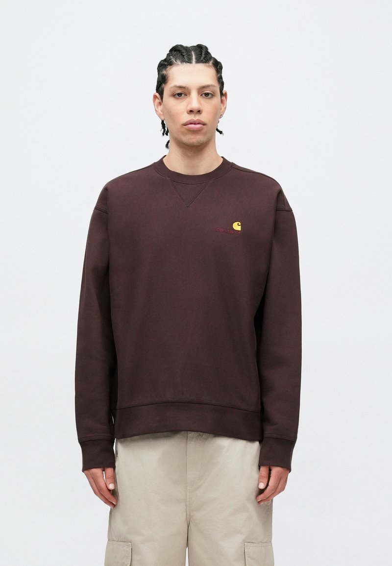 Braunes Fleece-Sweatshirt mit gerippten Bündchen und Saum, mit kleinem gelbem Logo auf der Brust, getragen über hellbeigen Hosen.