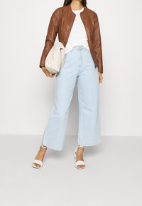 Giacca di pelle marrone, t-shirt bianca, jeans a gamba larga azzurri e décolleté bianche aperte. Accessorio: borsa a spalla beige con texture.