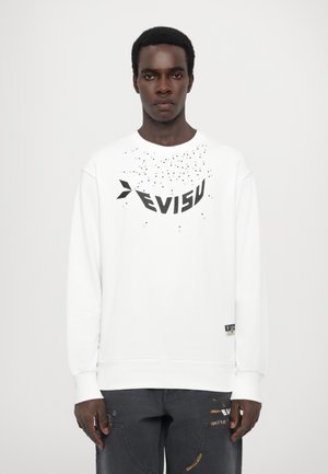 Hvit genser med svart "EVISU" logo og spredt prikkedesign. Lange ermer med ribbede mansjetter, kombinert med mørke cargo bukser.