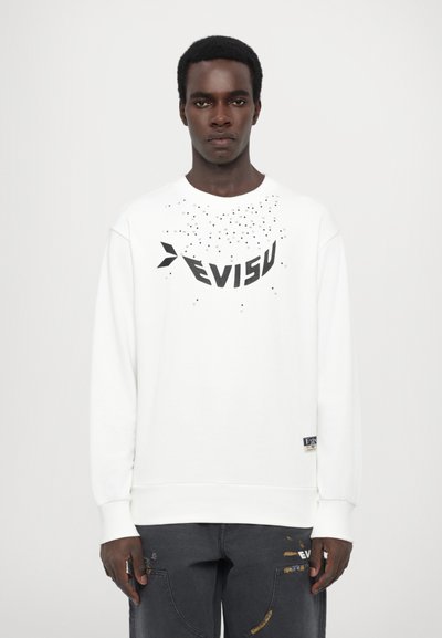 Weißer Pullover mit schwarzem "EVISU"-Logo und verstreutem Punktdesign. Lange Ärmel mit gerippten Bündchen, kombiniert mit dunklen Cargo-Hosen.