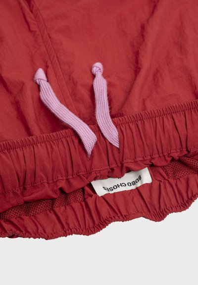 Ceinture élastique rouge avec cordons de serrage roses et étiquette partielle en tissu blanc affichant un texte noir sur un tissu léger.